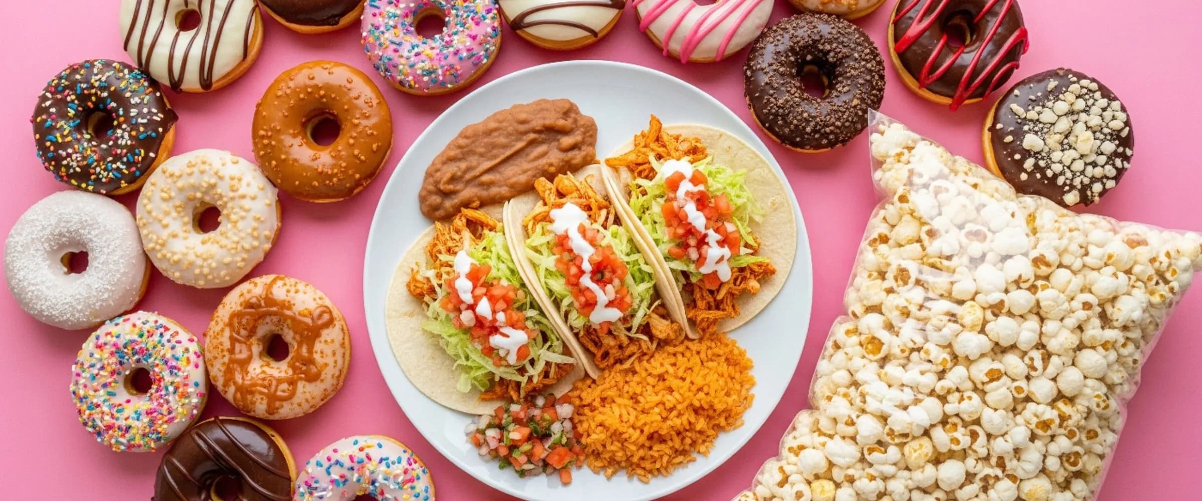 Glazed and sprinkled donuts, chicken tacos with rice and beans, and a bag of fresh popcorn on a pink table